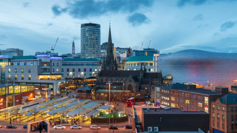 Cityscape of Birmingham at dusk featuring historic and modern architecture, including a church with a spire, shopping areas, and contemporary buildings like the Selfridges store. The image captures a vibrant urban atmosphere with street activity and a mix of old and new structures under a dramatic sky.