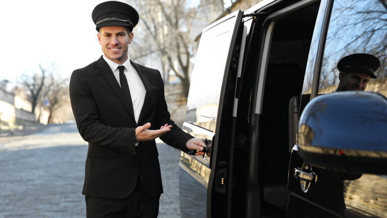 Professional chauffeur in a black suit and cap opening the door of a luxury vehicle, welcoming passengers on a sunny day.