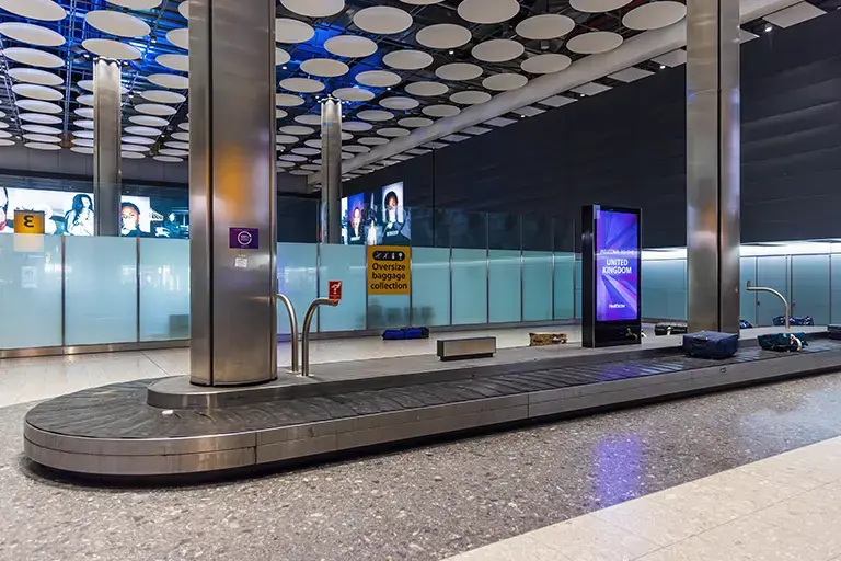 Baggage claim area at an airport featuring a luggage carousel, overhead signs for overseas baggage collection, and digital screens displaying information. The modern interior design includes a sleek metal structure and a high ceiling with circular light fixtures, providing a spacious and welcoming atmosphere for arriving travelers.