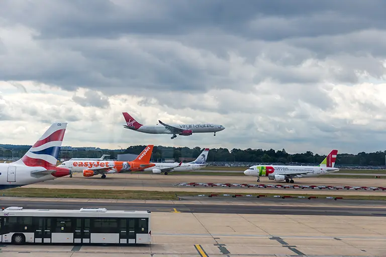 Airplanes from various airlines, including British Airways, easyJet, and TAP Air Portugal, parked on a runway, with a Virgin Atlantic flight preparing to land in the background under a cloudy sky. A bus is visible in the foreground, indicating airport activity.