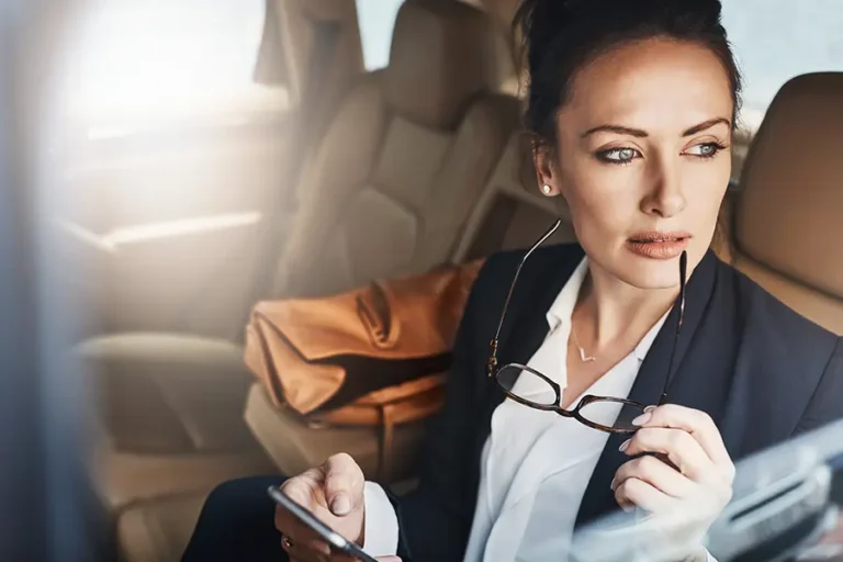 Businesswoman sitting in a car, holding glasses and a smartphone, looking thoughtfully out the window, dressed in a professional outfit with a brown handbag visible in the background.