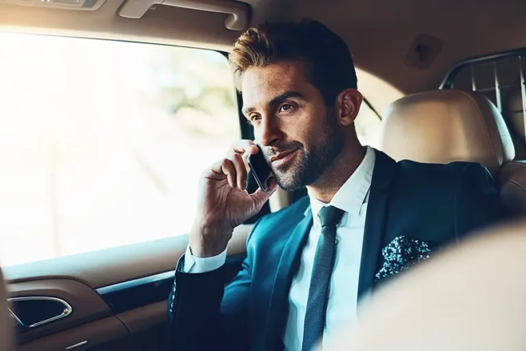 Businessman in a suit sitting in a luxury car, engaged in a phone conversation with a thoughtful expression. Natural light illuminates the interior, highlighting a modern and sophisticated atmosphere.