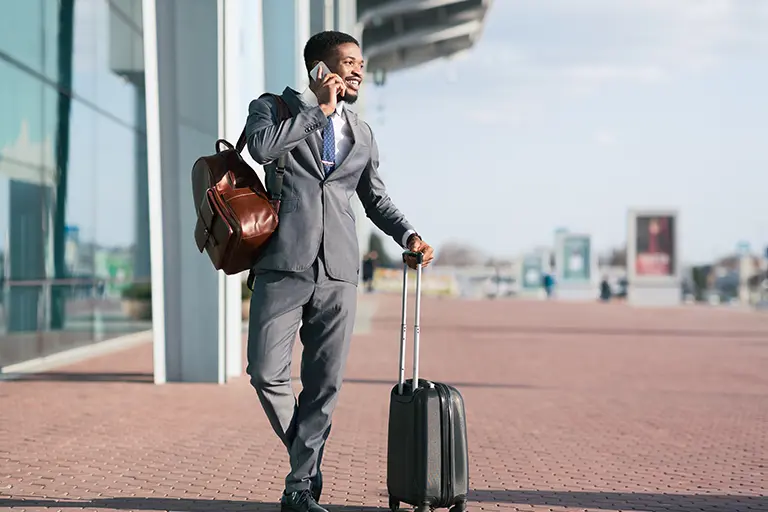 Businessman in a suit holding a suitcase and a leather bag, talking on the phone while walking at an airport terminal.