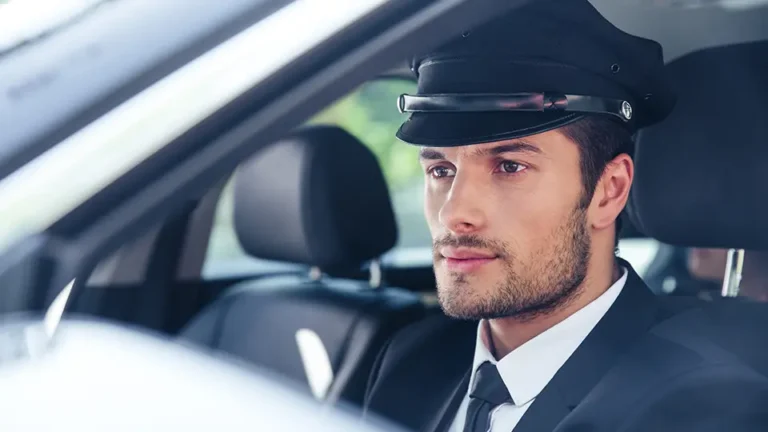Professional chauffeur in a black suit and cap, sitting in the driver's seat of a luxury vehicle, focused and ready to drive.