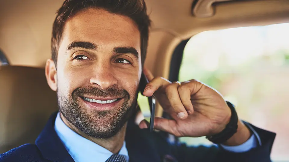 A smiling businessman in a suit talks on his smartphone while sitting in a car, exuding confidence and professionalism.