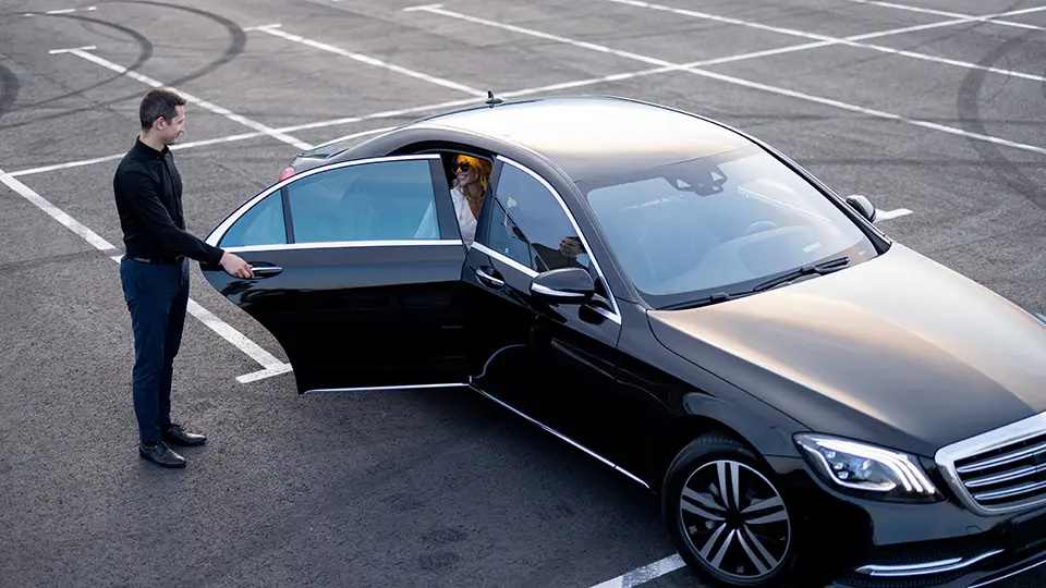 VIP arrivals chauffeur pickups Birmingham Airport - a man opening the passenger door of a black luxury car for a woman inside, set in an empty parking lot.