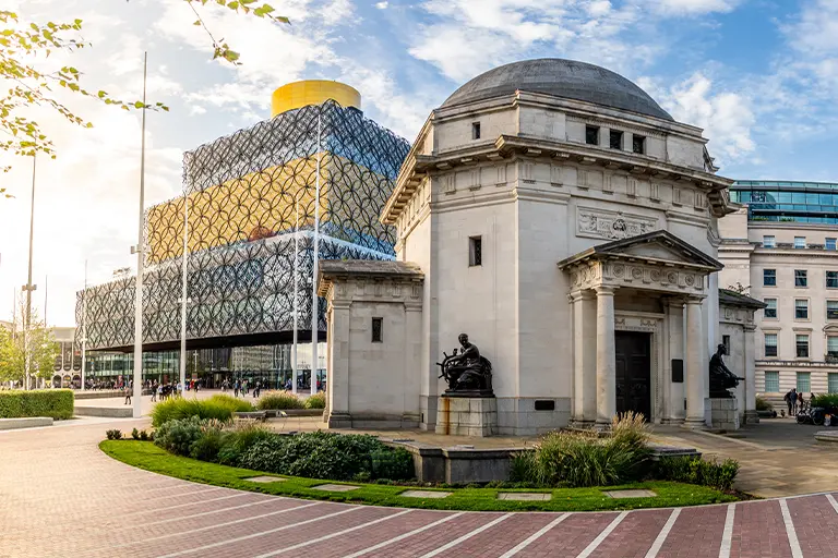 Chauffeur for special events - historic building in front of the modern Library of Birmingham, showcasing a blend of architectural styles with lush greenery and a clear blue sky.