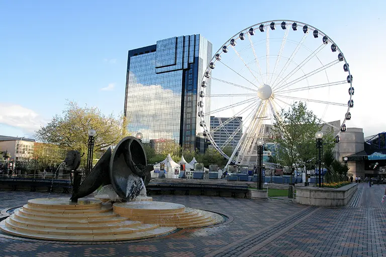 A scenic view of a city plaza featuring a decorative fountain and a large Ferris wheel in the background, surrounded by modern skyscrapers and greenery. The image captures the vibrant atmosphere of urban life and leisure activities.