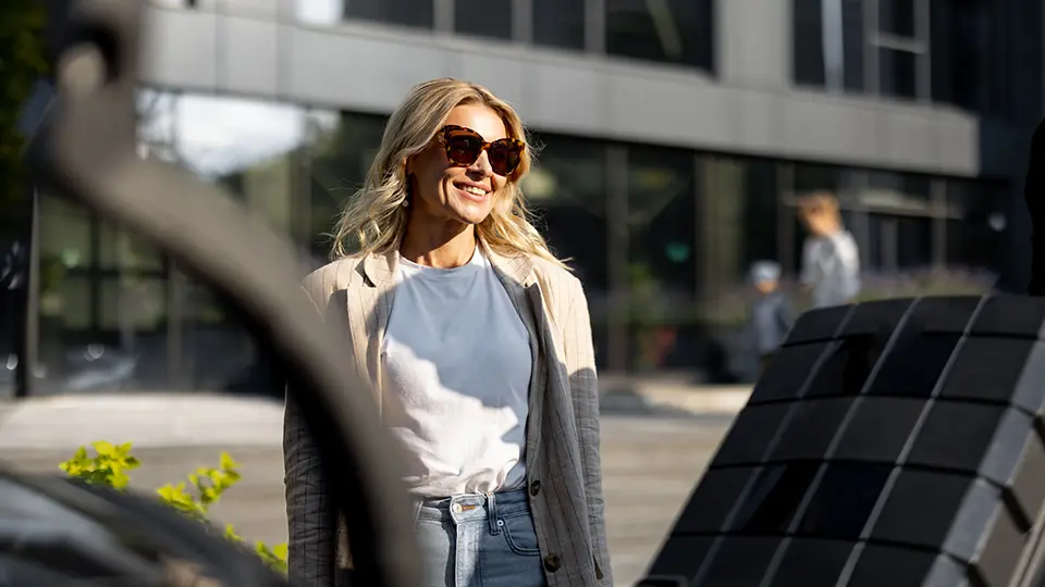 Smiling woman in stylish sunglasses stands outdoors in an urban setting, showcasing a modern building. She wears a light blazer over a casual top and jeans, reflecting a trendy, professional look in a bright, sunlit environment.