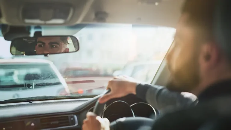 Man driving a car, focusing on the road ahead while checking the rearview mirror, with a sunny day illuminating the interior.