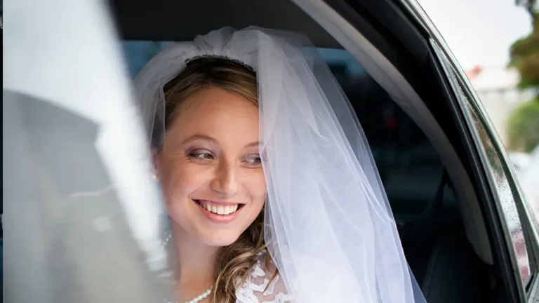 Bride smiling in a wedding dress and veil while sitting in a car, radiating joy and anticipation for her special day. Natural light enhances her happy expression.