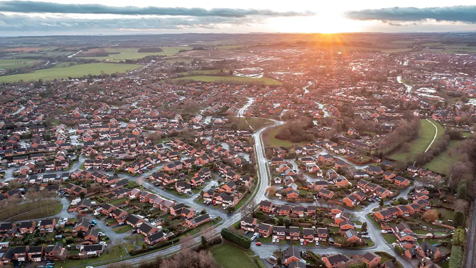 Aerial view of a suburban neighborhood at sunset, showcasing rows of houses with red roofs, winding streets, and surrounding green fields. The image captures the tranquility of residential life and the beauty of the landscape.