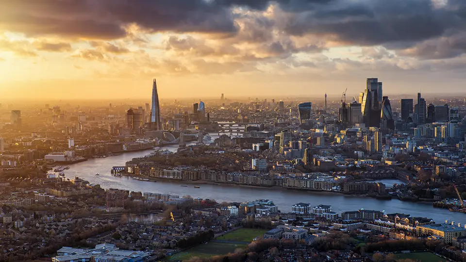 Aerial view of London at sunset, showcasing the River Thames winding through the city, with iconic skyscrapers like The Shard and the Gherkin illuminated by warm golden light. The skyline features a mix of modern architecture and residential areas, creating a stunning urban landscape.