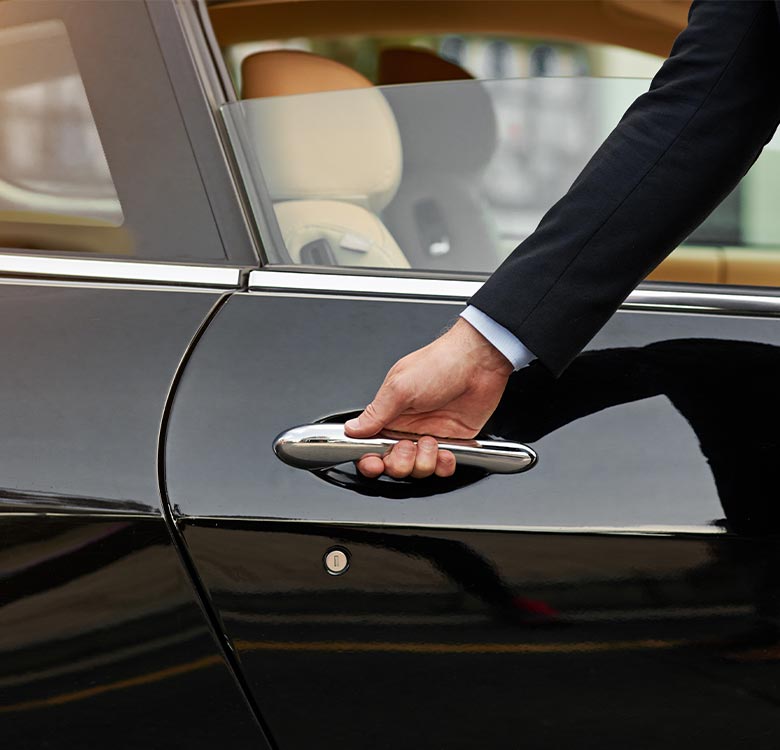 A businessman in a suit reaching for the door handle of a black luxury car, preparing to enter the vehicle. The image showcases elegance and professionalism in a corporate travel setting.