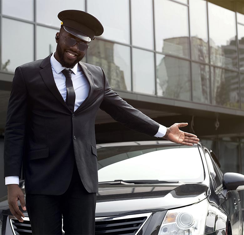 Chauffeur service Hagley - a professional chauffeur in a black suit and cap stands beside a luxury car, smiling and gesturing welcomingly in front of a modern building.