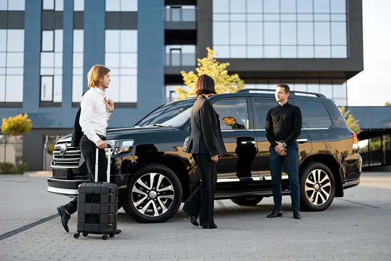 Business professionals meeting near a luxury black SUV, with one person holding a suitcase, in front of a modern building. The scene conveys business travel and professionalism.