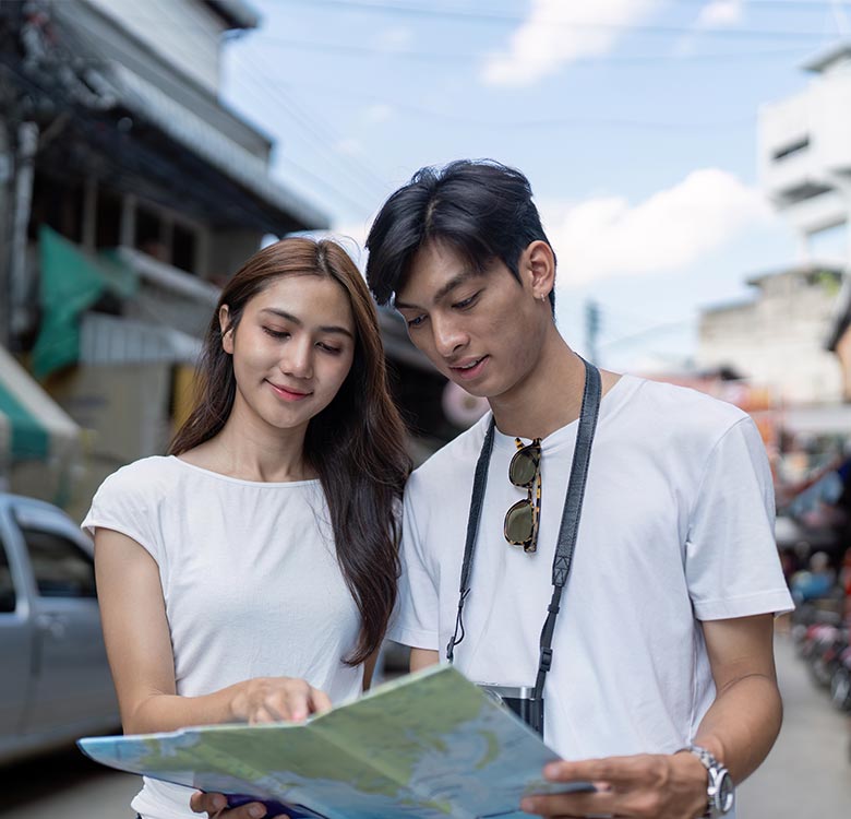 Young couple examining a map in a bustling outdoor setting, highlighting travel exploration and adventure. The man wears sunglasses and a camera strap, while the woman smiles, demonstrating a shared interest in discovering new places.