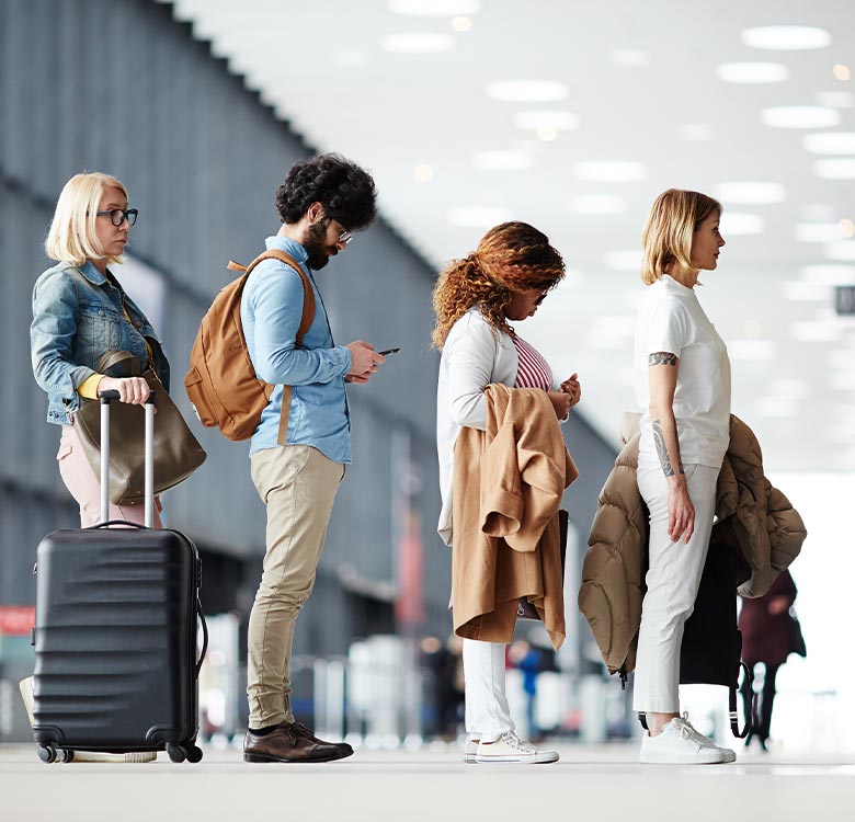 A diverse group of travelers waiting in line at an airport check-in counter, including a woman with glasses and a suitcase, a man checking his phone, and two women dressed in casual attire.
