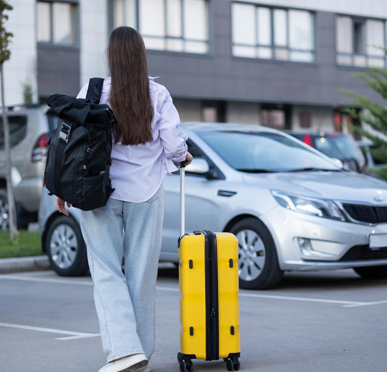 Young woman with long hair walking in a parking lot, carrying a black backpack and pulling a yellow suitcase, with cars and a modern building in the background. Ideal for travel and adventure-related content.