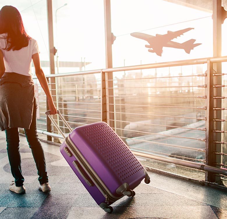 Airport transfers from Birmingham to Manchester - woman with purple suitcase walking through an airport terminal, with an airplane flying in the background as sunlight streams through the windows.