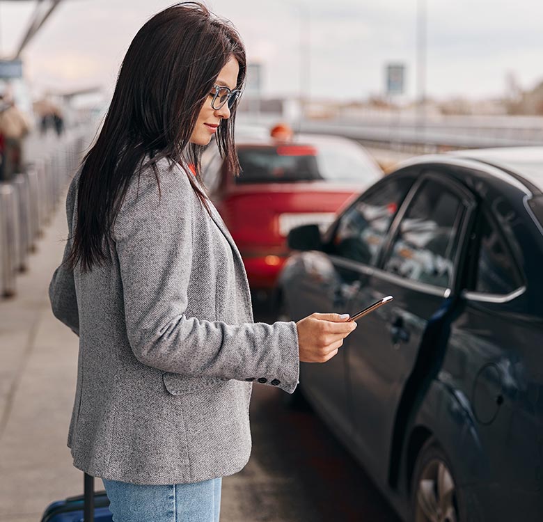 Woman in a gray blazer checking her phone while standing next to parked cars at an airport. She appears to be waiting or preparing for travel, indicating a busy travel environment.