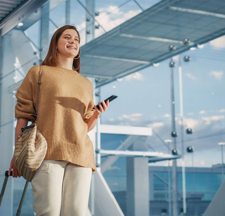 Woman smiling at the airport while holding a smartphone and standing next to a suitcase, dressed in a cozy sweater and casual pants against a modern glass backdrop.
