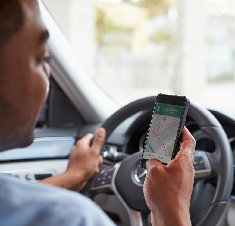 Driver using a smartphone for navigation while holding the steering wheel, showing a map with directions on the screen.