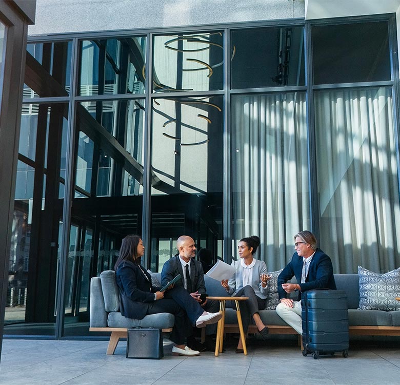 Business professionals engaging in a meeting in a modern, glass-walled lounge area, featuring stylish furniture and natural light, with a briefcase and luggage visible.