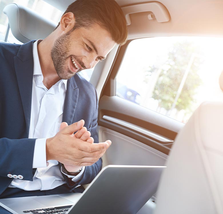 Businessman enjoying a successful moment while working on a laptop in the back seat of a car, dressed in a navy suit and smiling.