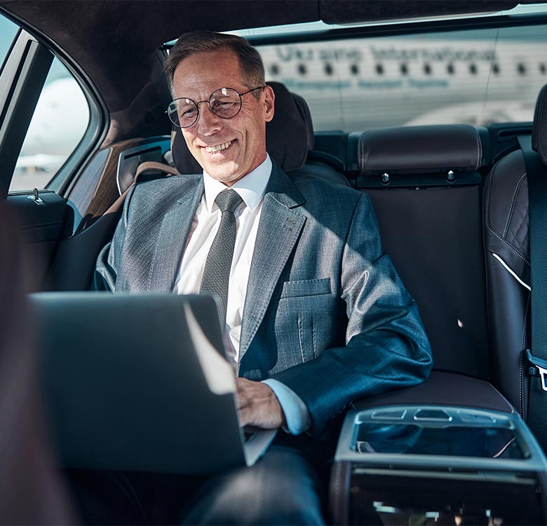 Chauffeur transfers from Birmingham to Manchester - businessman using a laptop in the back seat of a luxury car, smiling and dressed in a formal suit, with an airport terminal visible through the window.
