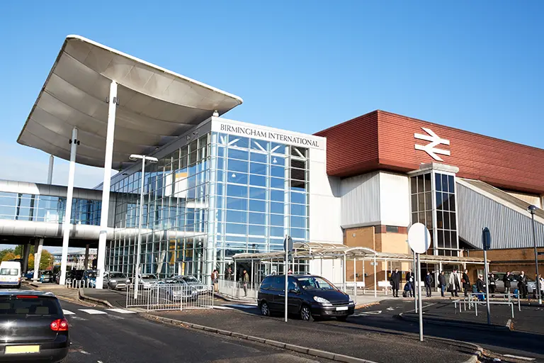 Birmingham International train station featuring modern glass architecture, with vehicles parked in front and passengers waiting outside under a clear blue sky.