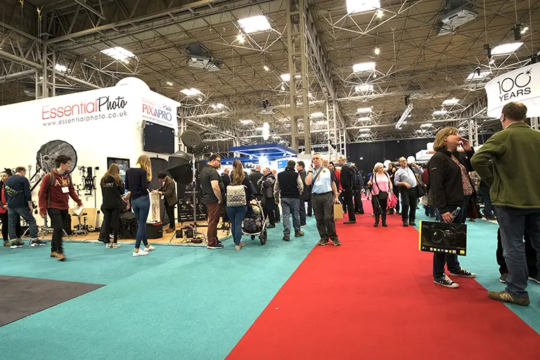 Crowded exhibition hall showcasing photography equipment at a trade show, with various booths including Essential Photo, attendees engaging in conversations, and displays of lighting and camera gear.