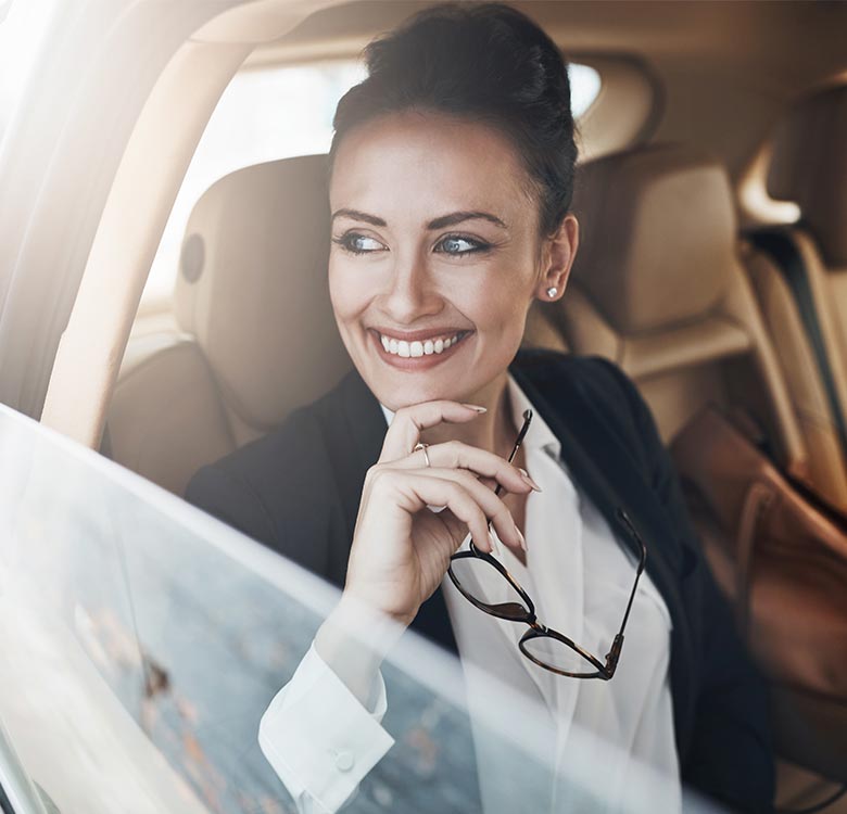Chauffeur service Oldbury - professional woman smiling in a luxury car, wearing a blazer and holding glasses, conveying confidence and success.