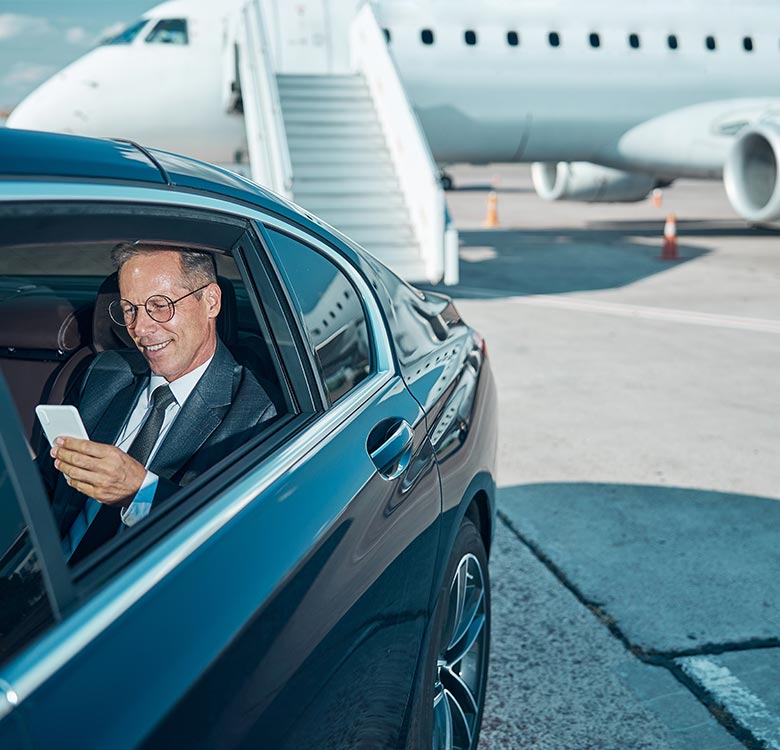 Businessman in a suit using a smartphone while seated in a luxury car at an airport, with an airplane and boarding stairs visible in the background.