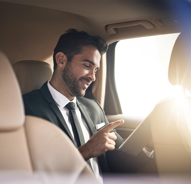 Businessman in a suit smiling and using a tablet inside a car, showcasing modern commuting and technology integration.