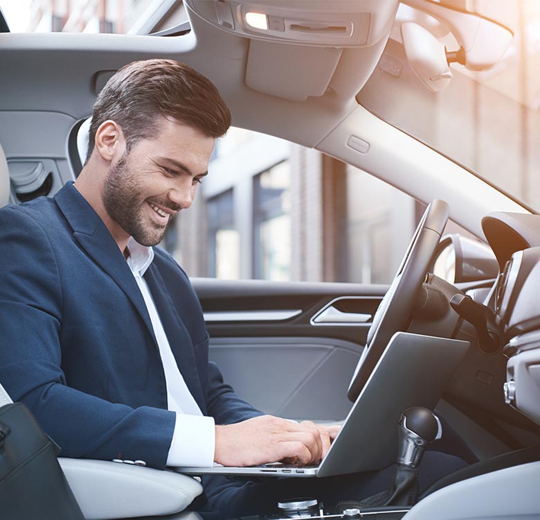 Book a reliable chauffeur for Birmingham Airport - businessman working on a laptop inside a modern car, smiling as he focuses on his tasks, with a cityscape visible through the windows.