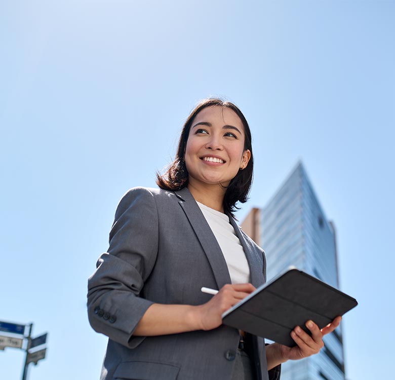 Smiling businesswoman in a gray suit holding a tablet, standing outdoors with a clear blue sky and modern buildings in the background, representing professionalism and technology in the workplace.