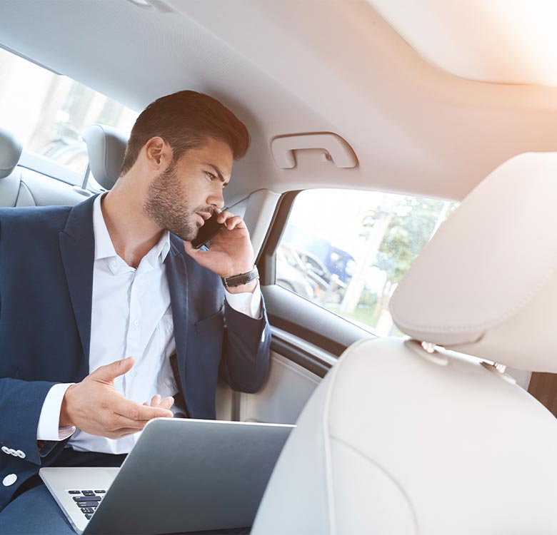 Businessman in a suit sitting in the backseat of a car, using a laptop and talking on a phone, illustrating mobile work and productivity during travel.