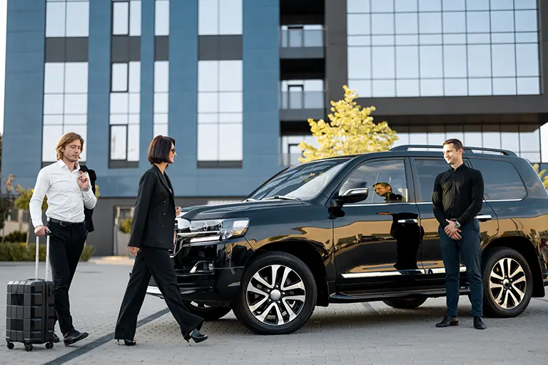 Business professionals arriving at a modern building, with a woman in a black suit walking toward a black luxury SUV while a man waits nearby. A man with a suitcase appears to be accompanying her, indicating a luxury transportation service or airport transfer setting.