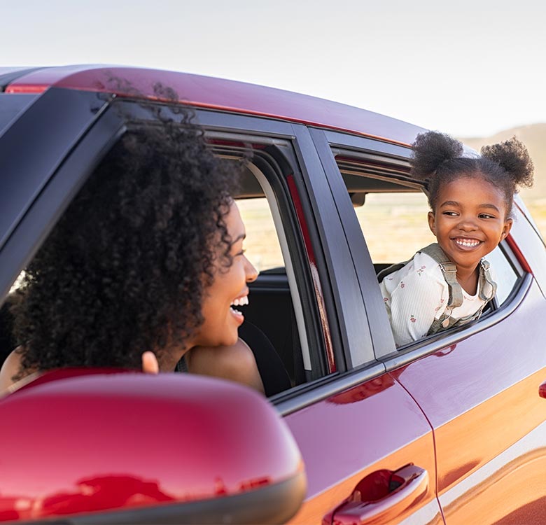A joyful mother and daughter smiling and interacting from a red car window during a sunny day, symbolizing family bonding and adventure in travel.