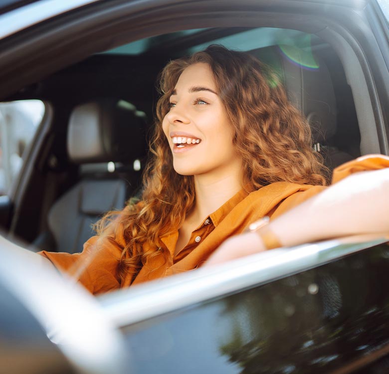 Smiling woman with curly hair leaning out of a car window on a sunny day, wearing a casual orange shirt, radiating a joyful and carefree vibe.