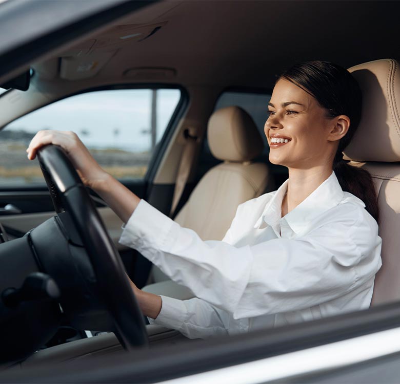 Smiling woman driving a car, wearing a white shirt, focused on the road with a modern interior in the background, showcasing an enjoyable driving experience.
