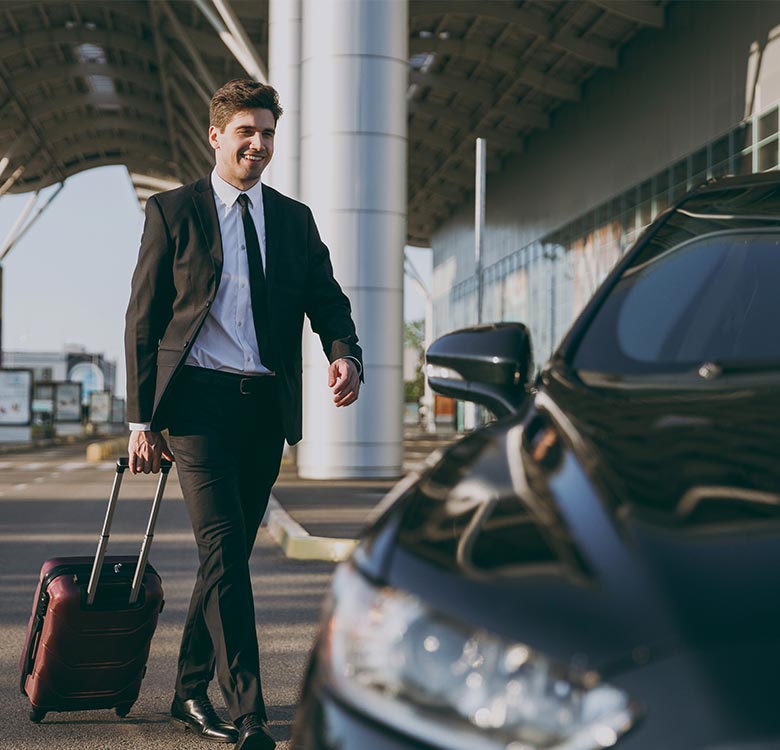 Happy businessman in a suit walking with a suitcase towards a car in an airport setting, ready for travel or a business trip.