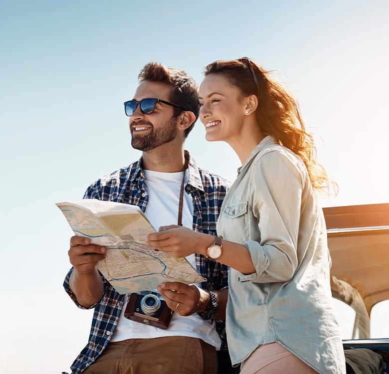 Young couple exploring travel destinations, smiling and looking at a map, with a camera hanging around the man's neck, set against a bright, sunny sky.