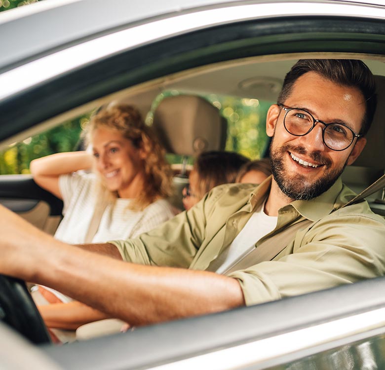 Happy man driving a car with family in the background, enjoying a road trip on a sunny day.