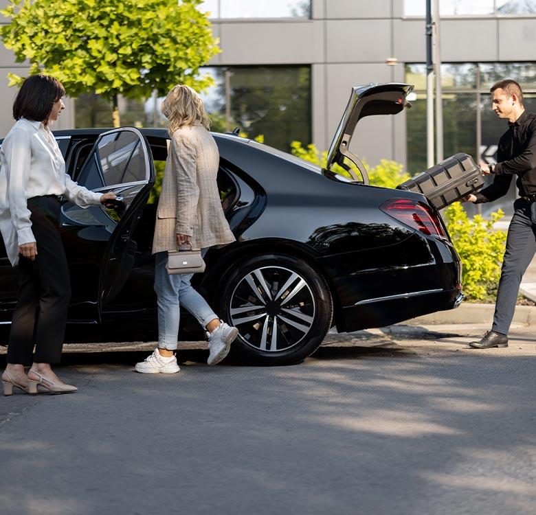 Women entering a luxury black car while a man loads luggage into the trunk, set in an urban environment with greenery and modern architecture in the background.