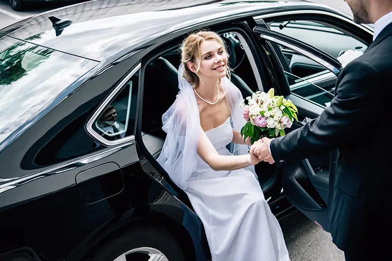 Bride in a white wedding dress and veil happily exiting a black car while shaking hands with a man, holding a bouquet of flowers, capturing a joyful wedding moment.