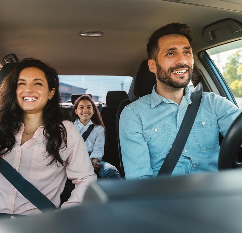 Happy family enjoying a road trip, with a woman and a man driving, and a girl smiling in the backseat, all wearing seatbelts.