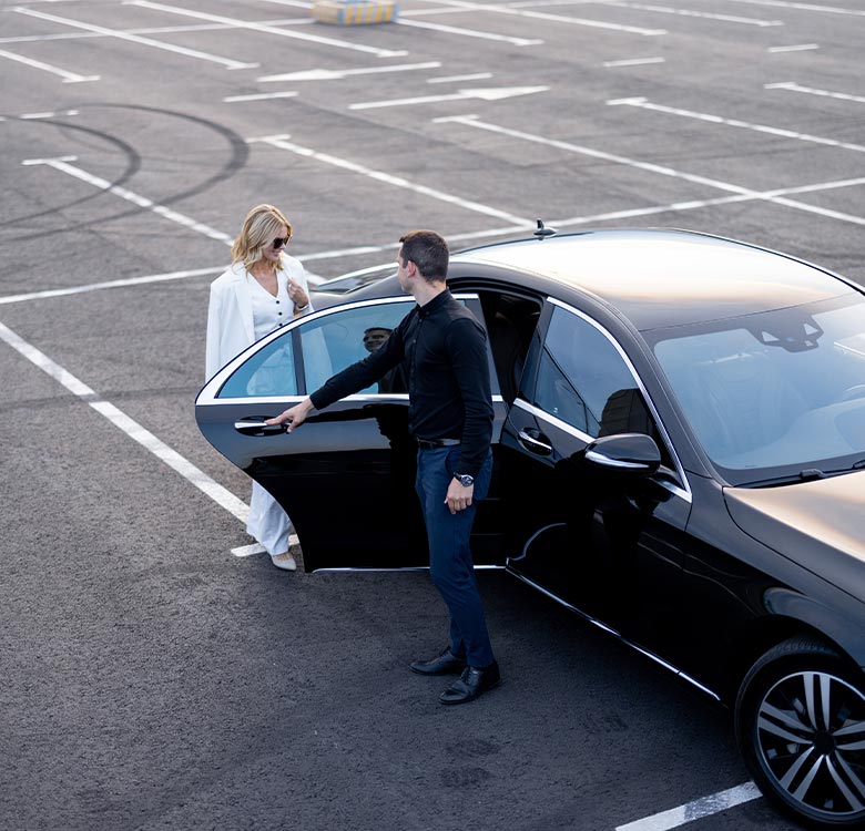 A stylish woman in a white outfit is stepping out of a luxury black car, while a well-dressed man in a black shirt holds the car door open, showcasing elegance and sophistication in a modern parking lot setting.