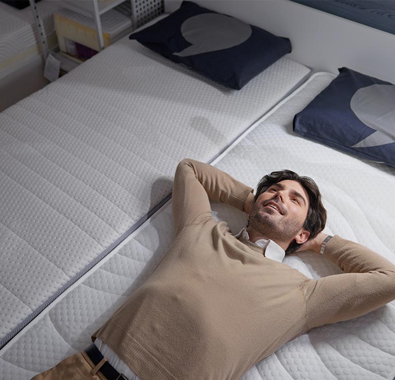 A man relaxing on a mattress in a showroom, showcasing the comfort and quality of modern bedding accessories, with stylish pillows in the background.